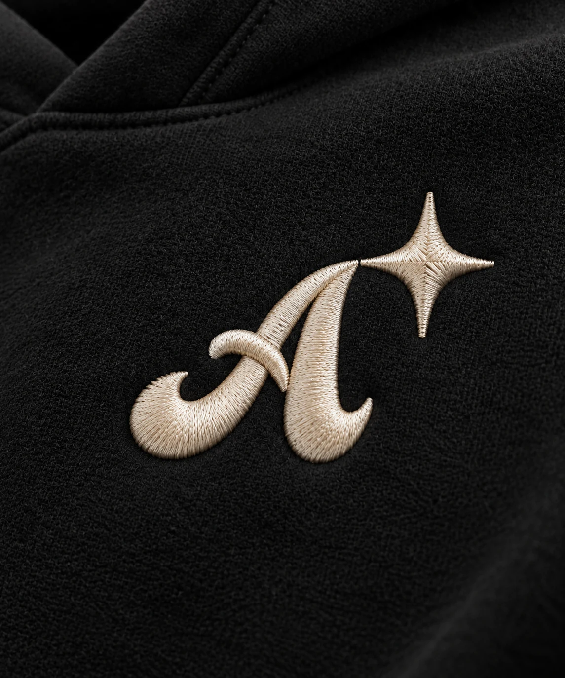 Macro close-up of the Ambifest A and four-point star in raised warm ivory satin stitch on black fleece.